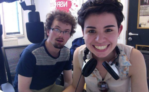 Bridget in the radio studio with headphones around her neck, smiling to camera with a fellow student.