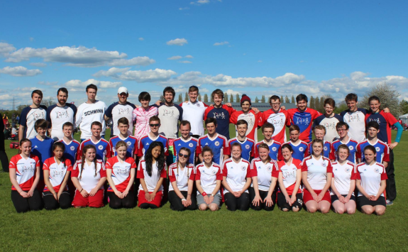 A group of student and alumni frisbee players posing for a team photo on grass with a blue sky above