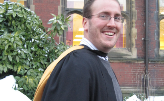 Dan smiling on campus on graduation day wearing a black robe