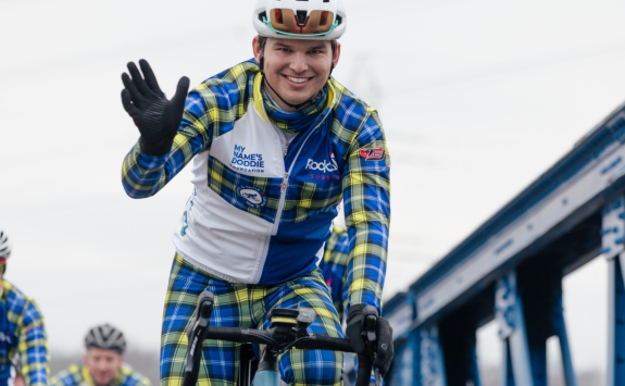 Fergus in tartan cycling gear riding over a bridge and waving to camera