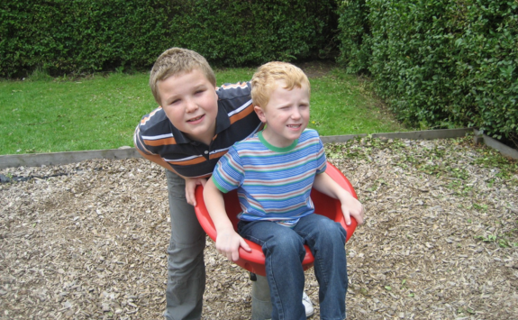 Peter and Daniel Hayton as children. Daniel sits in a spinning seat in a playpark while Daniel leans over him smiling.