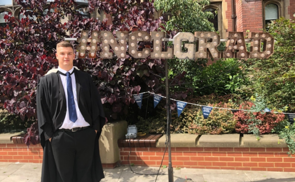 James on campus during graduations. He wears a black gown and blue tie, and is stood with his hands in his pockets in front of a NCL Grad light display.