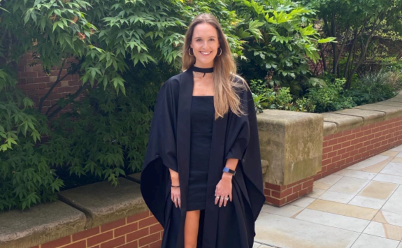 Lily in a black graduation gown and black dress posing in front of green foliage on campus
