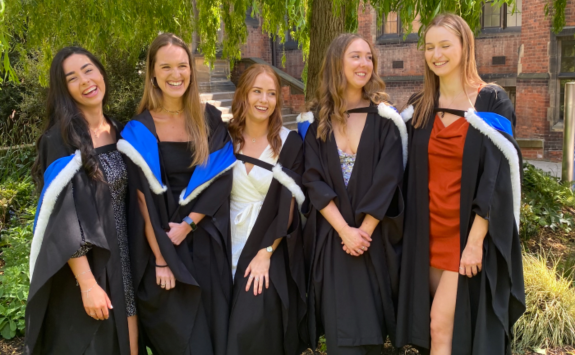 Lily and friends donning graduation gowns smiling under the willow tree on campus