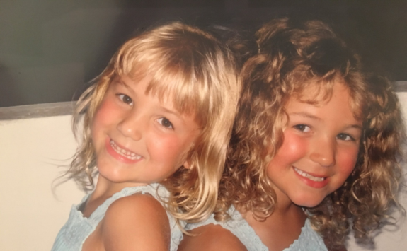 Jessica and Katie as children on holiday, smiling while sat on a bench with their backs to each other.