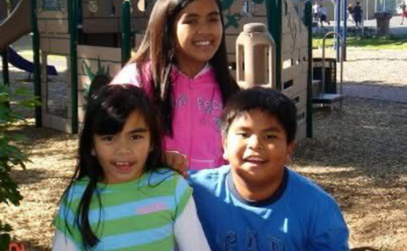 Three Sembranos siblings as children stood smiling in a play park