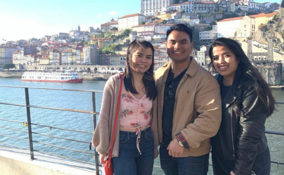Three Sembranos siblings as adults smiling in front of a river with a grand building in the background.