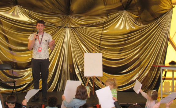 Will stood on stage in front of a gold curtain speaking to a crowd of children