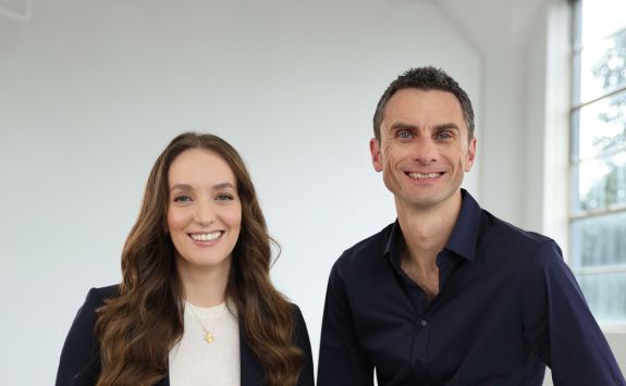 Alex and Justin are looking toward the camera. Alex is on the left. She has long dark hair and is wearing a white top and dark jacket. Justin is on the right. He has short, dark hair and is wearing a black, open-necked shirt.