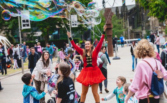 A science busker is creating large bubbles as part of an open air demonstration. She is wearing a red dress and is surrounded by young children who are watching the show.