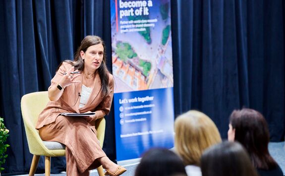 Estelle Blanks, Director of Newcastle Innovations, is sitting in a yellow fabric chair on a stage with a dark blue, curtained background. She is talking to the audience. She has long, dark hair and is wearing a dusky blush suit with a white top.