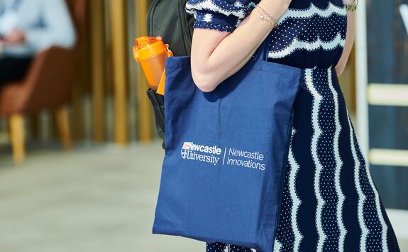 A woman in a dress is seen standing with a blue tote bag hanging from her shoulder. The bag carries the Newcastle University logo and the Newcastle Innovations word mark.
