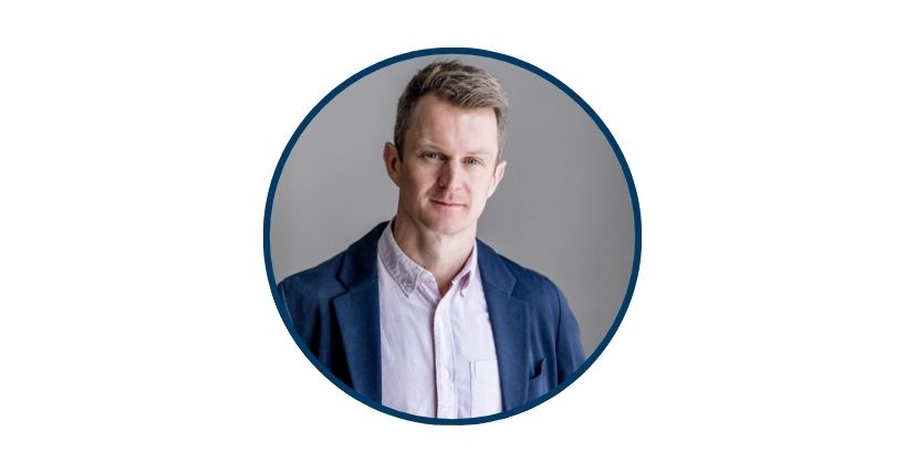 Dr Oliver Shannon is a white male with short, fair hair. He is pictured wearing a mauve coloured shirt and blue jacket.
