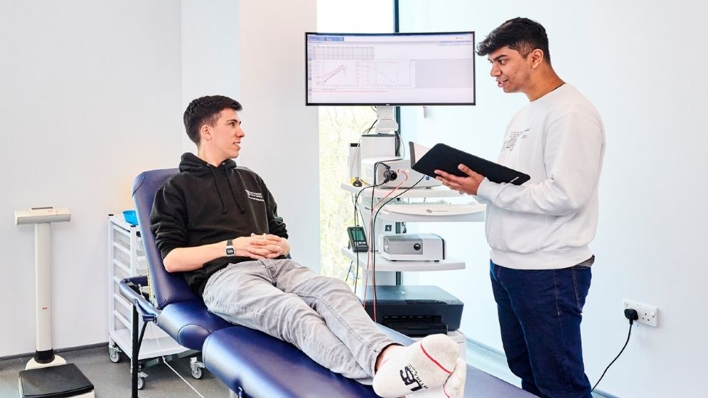 A male participant is undergoing metabolic assessment in a clinical research facility while a make researcher records measurements using specialist monitoring equipment and data displayed on a computer screen.