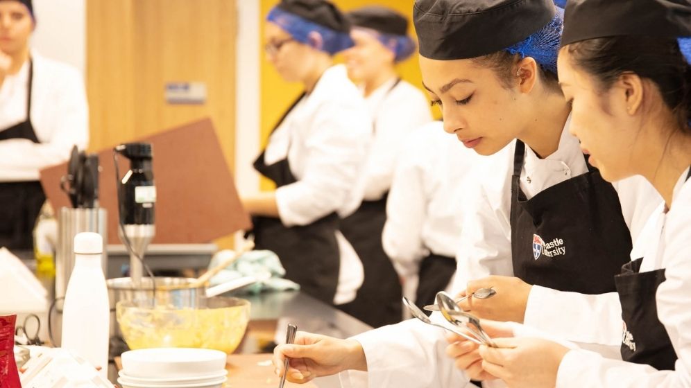 Participants using a specialist nutrition training kitchen, preparing and assessing food products in a practical food science and nutrition environment. they are wearing chef whites, black hats and black aprons.