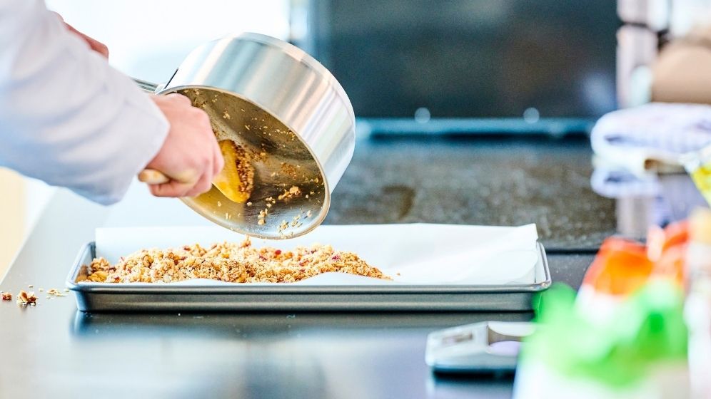 Food is being poured out of a pan and into a baking tray lined with greaseproof paper.