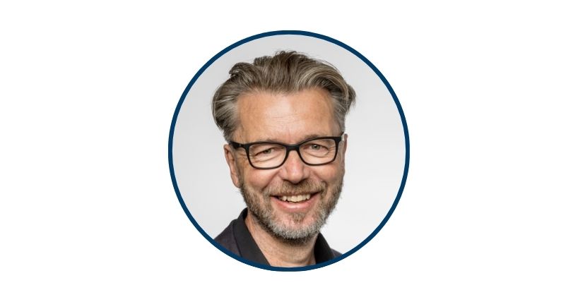 Prof Bernard Corfe is a white mail with dark, greying hair and beard. He also wears glasses.