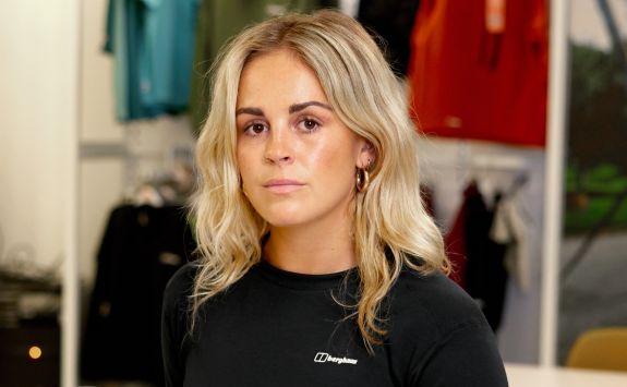 A person with long blonde hair wearing a black Berghaus shirt stands indoors in front of clothing racks displaying green and red shirts.