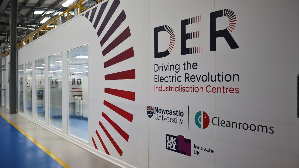 Wall inside a modern industrial facility featuring the 'Driving the Electric Revolution Industrialisation Centres' logo and graphic design, with visible cleanroom equipment through adjacent windows. Logos for Newcastle University, Cleanrooms, and Innovate UK are also displayed.
