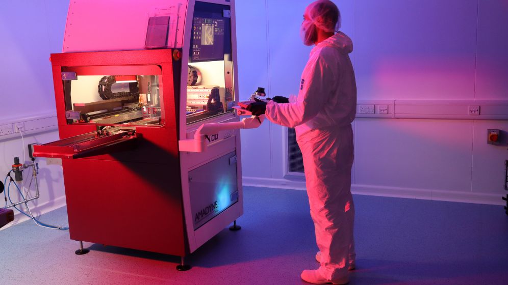 Operator in full cleanroom protective gear working with a red and white machine in a high-tech cleanroom environment, illuminated by blue and pink lighting.