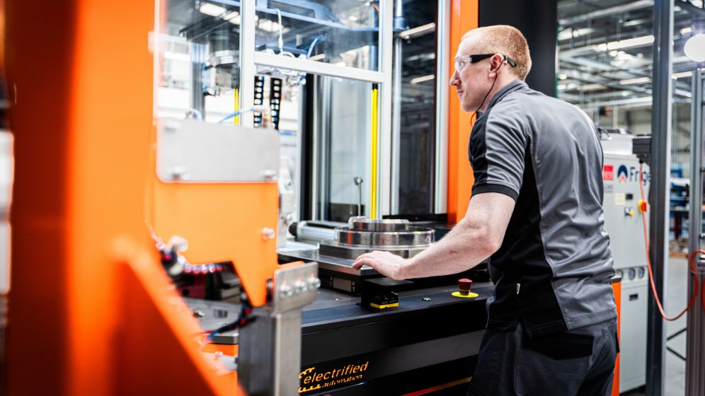 Technician in safety glasses and grey uniform operating orange and black machinery labeled 'electrified' in a well-lit industrial workspace.