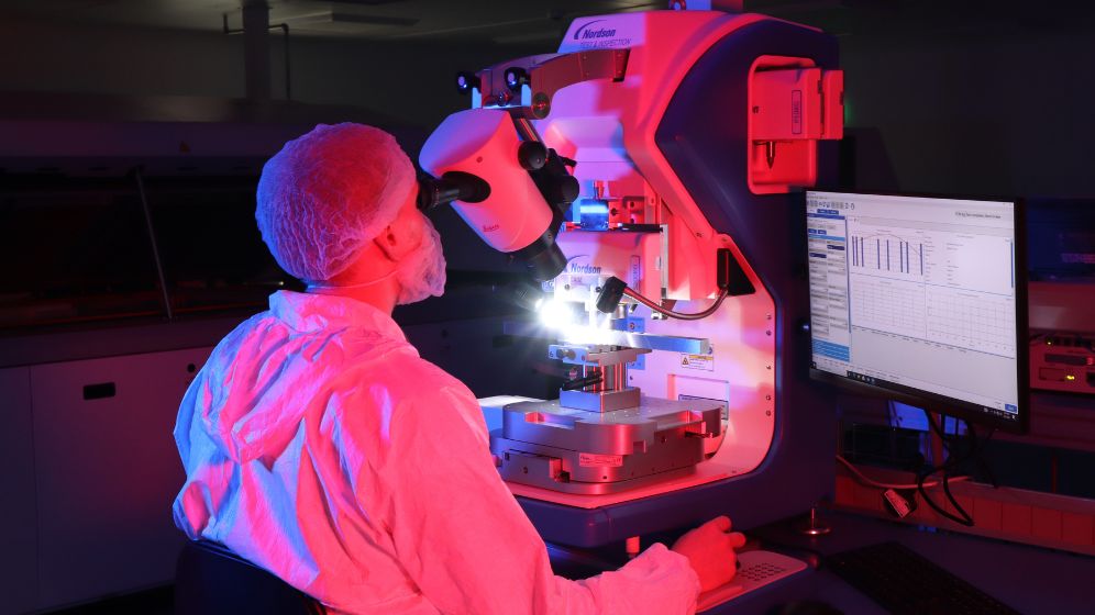 Technician in protective clothing operating a microscope-like machine under bright light in a dimly lit lab with blue and red lighting; data charts visible on adjacent monitor.