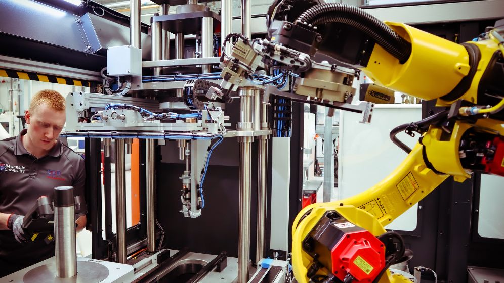 Technician operating a yellow robotic arm in a high-tech manufacturing environment, inspecting a cylindrical metal component on the machine's platform.