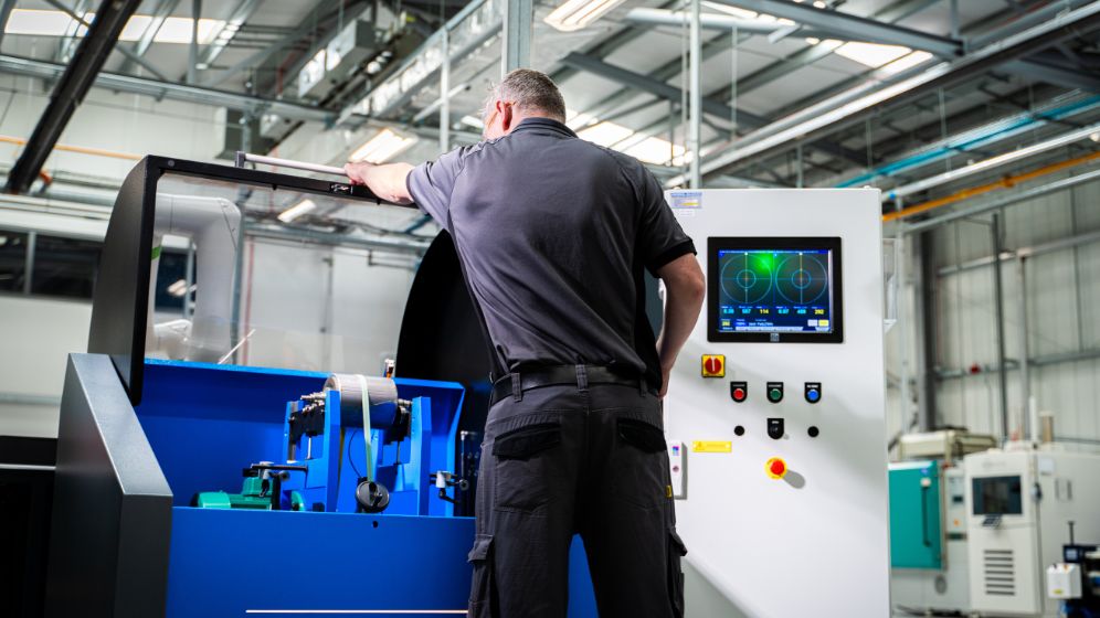 Operator working with a large industrial machine featuring a blue interior and control panel, set within a factory environment with visible beams, pipes, and equipment.