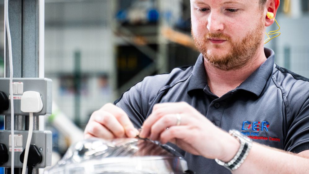 Technician wearing a DER-branded polo shirt and ear protection working on equipment in an industrial setting, with blurred machinery in the background.