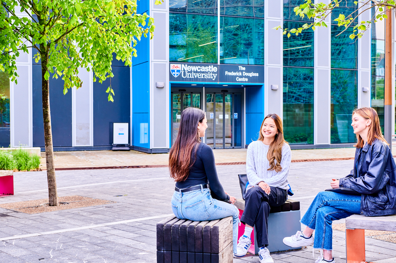 Three women sitting next to each other on a bench outside of the Frederick Douglass Centre building, engaged in conversation.