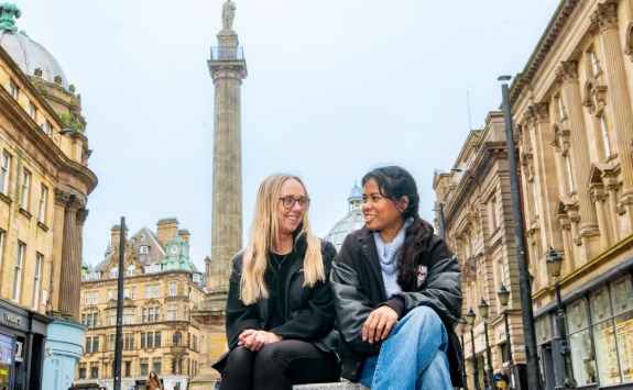 Two women sitting next to each other on a bench in Newcastle city centre, engaged in conversation. Behind them we can see Grey