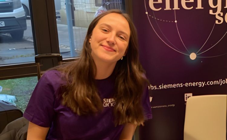 NUBS student Alice in a purple Siemens Energy T-shirt smiles while sitting at an event stand, with a Siemens Energy display banner behind her.