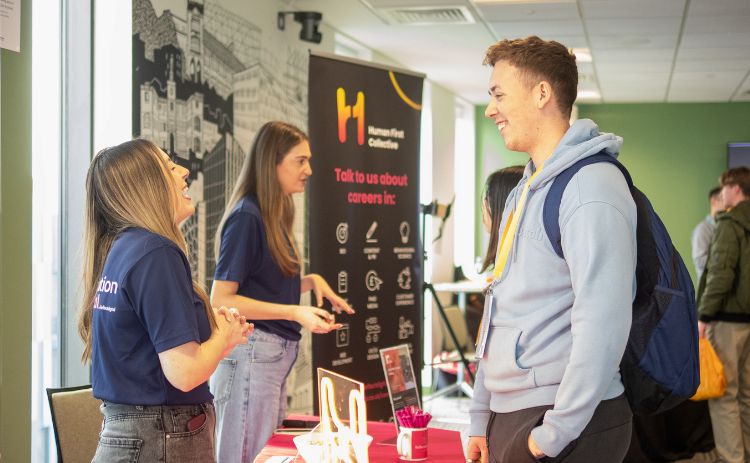 A student chats with two representatives at a careers stand during the Aspiration Digital 2025 event, with display banners behind them.