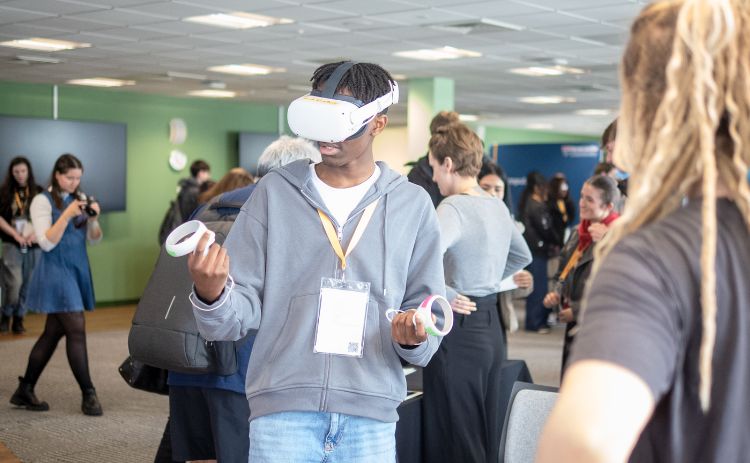 A young participant uses a virtual reality headset and controllers during an interactive session at the Aspiration Digital 2025 event, with others watching in the background.