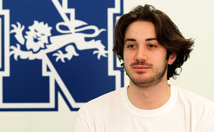 Austin Sacks wearing a white T-shirt in front of a wall displaying the Newcastle University sports logo