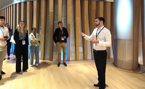 NUBS Alumnus Cameron Bateman leading a tour at Bloomberg, speaking to a diverse group of students in a modern office environment with wooden panel walls and bright lighting. Students listen attentively while standing in a semi-circle around the speaker.	