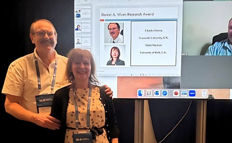 Charles Harvey and Mairi Maclean stand smiling beside a screen showing a slide for the Daniel A. Wren Research Award, with their names and photos displayed. A virtual participant is visible on the right.