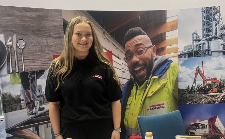 NUBS student Eleanor wearing an EGGER-branded black polo shirt stands smiling in front of a colourful EGGER display booth featuring images of employees and industrial scenes.