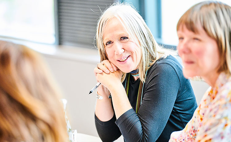 A light-skinned woman with long blonde hair smiling and holding a pen, engaged in a classroom discussion during an executive education session. Two other women, also light-skinned, sit beside her, partially visible. The background shows a bright, well-lit room