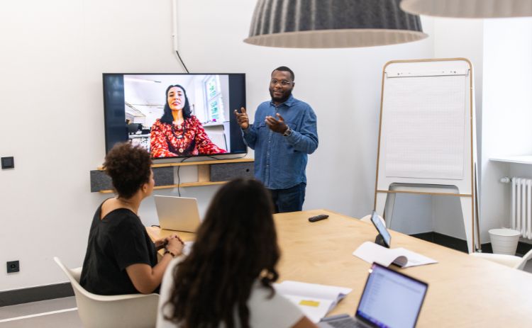 Presenter speaking to colleagues in a meeting room while a remote participant appears on the large screen; laptops and notes are visible on the table.