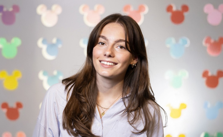 NUBS student Jess smiles at the camera, standing in front of a soft-focus wall covered with colourful Mickey-shaped icons.