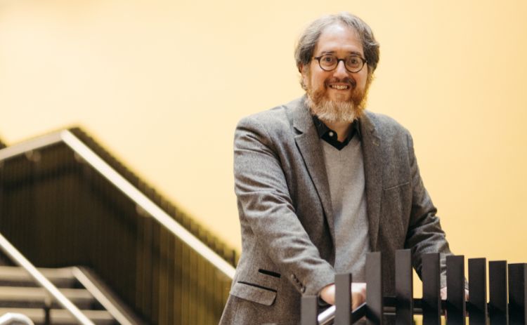 Professor Jonathan Sapsed stands smiling beside a black bannister, wearing a grey blazer and jumper, with a staircase and yellow wall in the background.