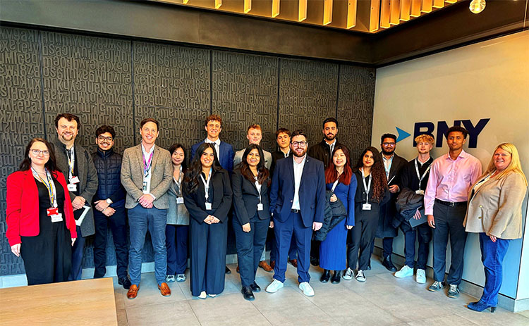  students and staff standing together at the BNY office, dressed in business attire, posing in front of a textured black wall with the BNY logo.