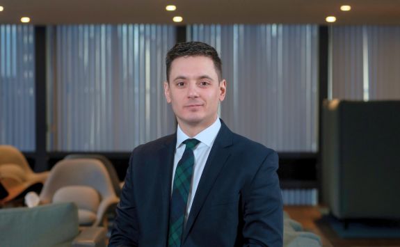 Marco Concina in a dark suit and green tartan tie stands in a modern office lounge. Vertical blinds and soft lighting are visible in the background.