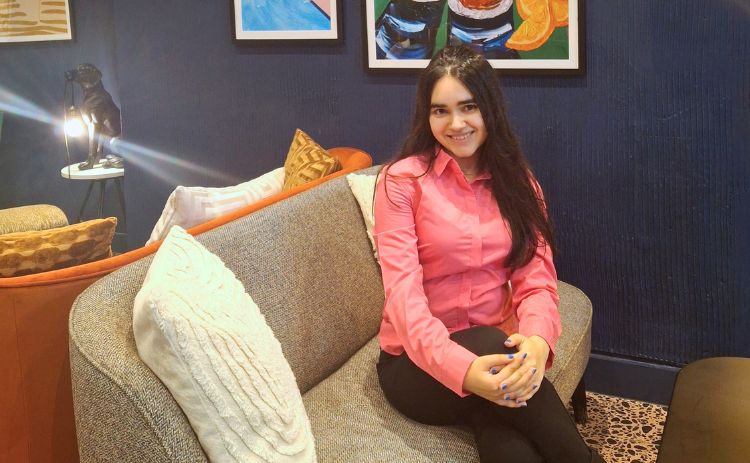 Mehek wearing a bright pink shirt and black trousers sits on a grey sofa in a stylish lounge area with patterned carpet, navy walls, framed artwork, and decorative cushions.