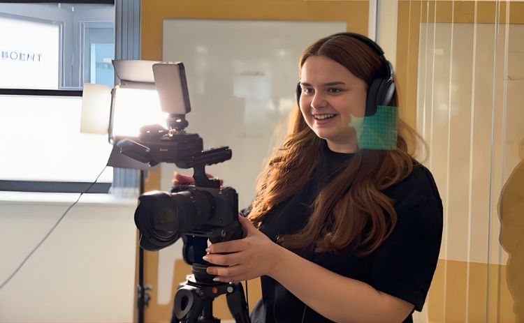 NUBS student Molly wearing headphones operates a professional video camera on a tripod, smiling as she films in a brightly lit room.