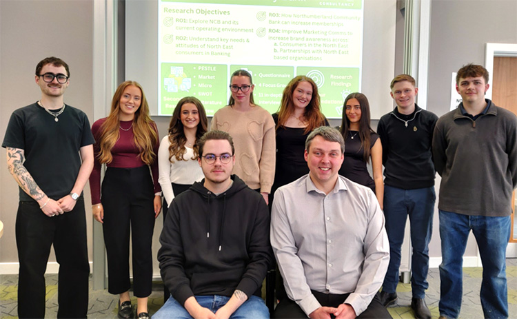 Eight students stand smiling behind two seated Northumberland Community Bank staff members. They’re posing in front of a presentation slide showing research objectives and findings related to the bank.