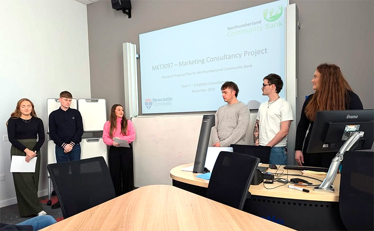 A group of students stands at the front of a classroom presenting their marketing consultancy project for Northumberland Community Bank, with a slide displayed on the screen behind them. 