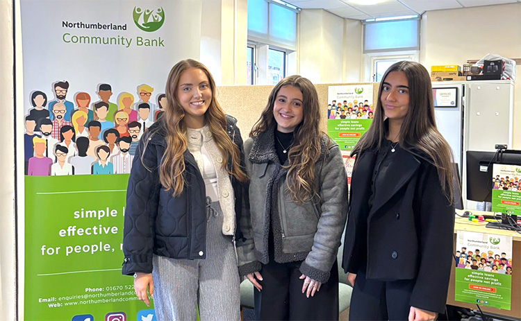 Three young women stand smiling in front of banners for Northumberland Community Bank. The banners feature a diverse illustrated crowd and text promoting the bank as "simple, effective for people." Office setting with posters and furniture visible.