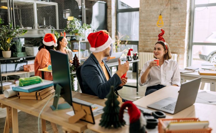A group of colleagues wearing festive hats and chatting around desks in an office decorated for Christmas.